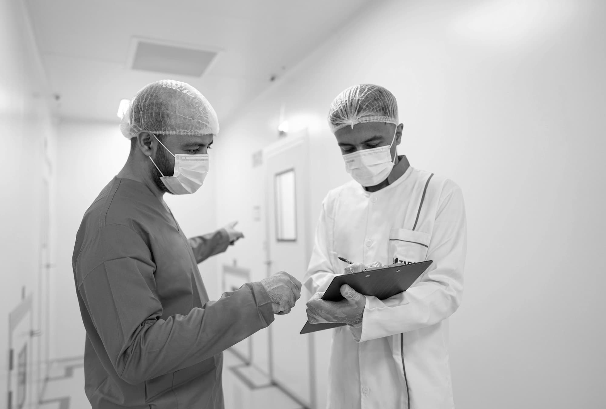Two clinicians in masks and hair covers reviewing a clipboard in a hospital corridor